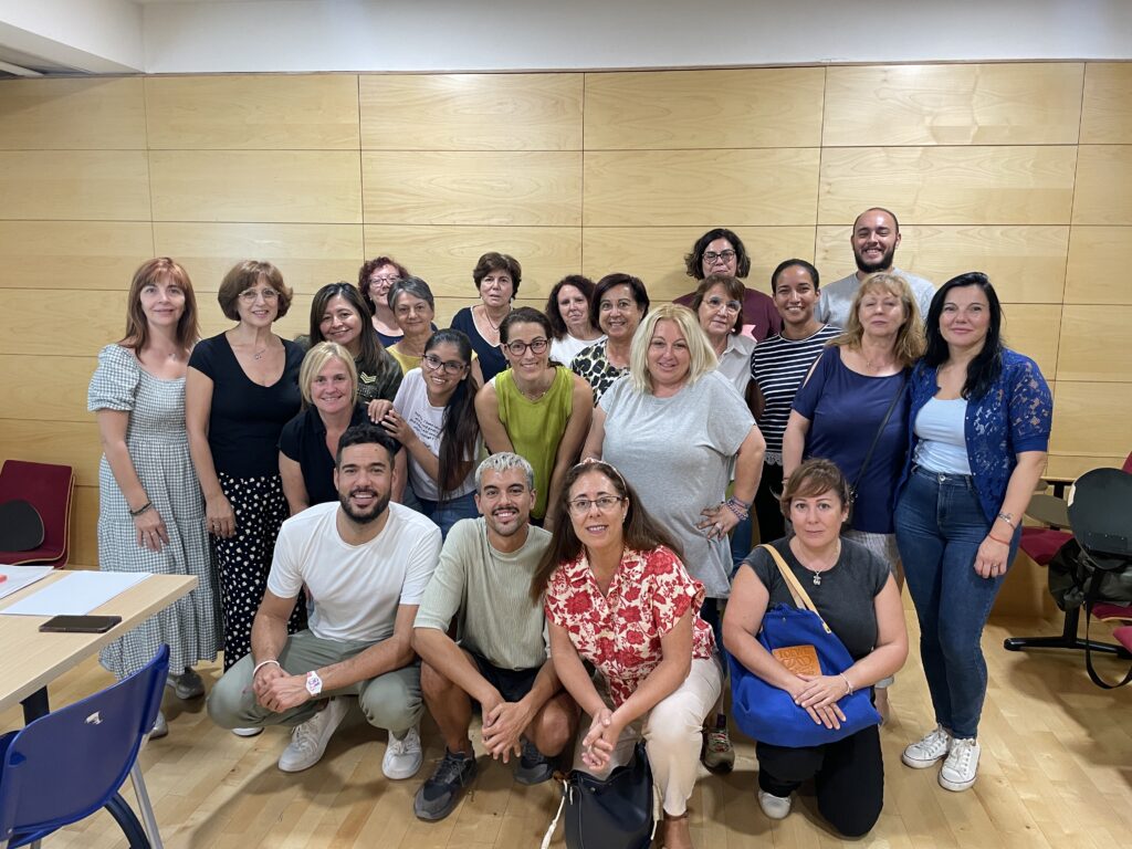 En la fotografía se muestra un grupo diverso de personas sonriendo y posando para la cámara en una sala luminosa con paredes revestidas de madera clara. Las personas están de pie y sentadas, y parecen haberse reunido para una ocasión especial o un evento. Las vestimentas son variadas, desde atuendos casuales hasta más formales. El grupo transmite una sensación de camaradería y alegría. La disposición de las personas en diferentes niveles, tanto de pie como sentadas, añade dinamismo a la imagen. En la parte inferior de la foto, hay sillas azules y una mesa con papeles y otros objetos sobre ella, lo que sugiere que la reunión pudo haber tenido un propósito educativo o profesional. La combinación de expresiones felices y el ambiente cálido de la sala hacen que la foto transmita un sentimiento de comunidad y colaboración