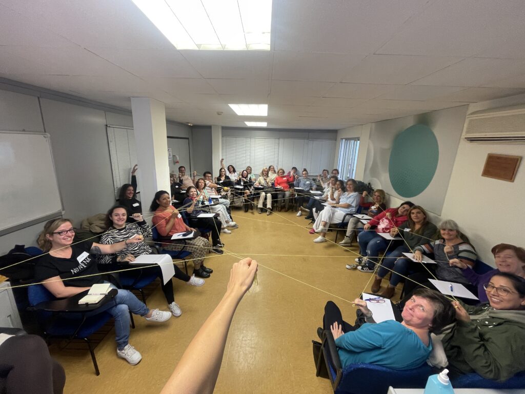 La imagen muestra un aula luminosa con paredes de color claro. En la sala, un grupo de personas se sienta en sillas dispuestas en hileras. Todas parecen estar de buen humor, algunas de ellas posan y sonríen a la cámara, mientras que otras levantan la mano o hacen gestos amistosos. Varias personas llevan ropa casual, y algunas tienen en sus manos cuadernos o papeles, sugiriendo un ambiente de aprendizaje o formación. En la parte frontal del aula, hay una pizarra blanca, y se pueden ver las manos de alguien, probablemente del fotógrafo o de un orador, extendidas hacia la audiencia. En general, la imagen transmite una atmósfera relajada y alegre, de camaradería y entusiasmo.